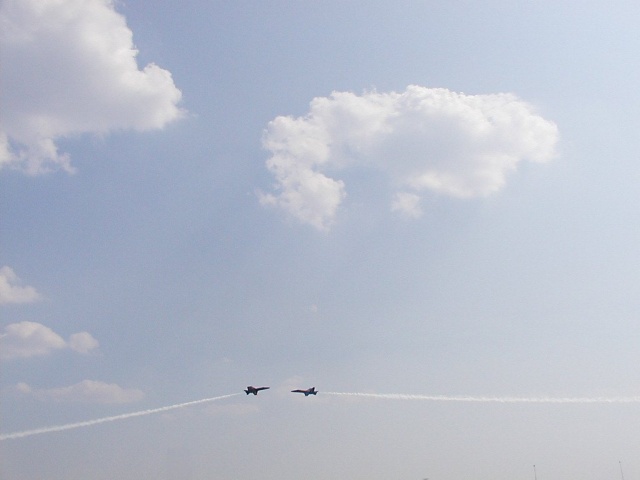 Two fighter jets fly across a cloudy sky, leaving contrails