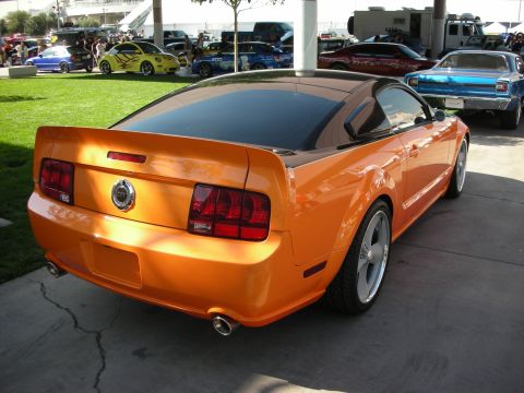 1998 Ford Crown Victoria - orange Ford Mustang coupe with polished wheels is parked on pavement, shown from the rear three-quarter among other show cars.