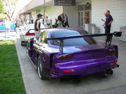 1998 Ford Crown Victoria - purple wide-body sports car with a tall rear wing and aggressive aero sits on a concourse near a Concourse C signage.