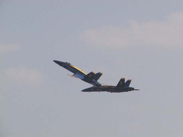 Two Blue Angels jets flying in formation against a cloudy sky. One jet is above and slightly ahead of the other