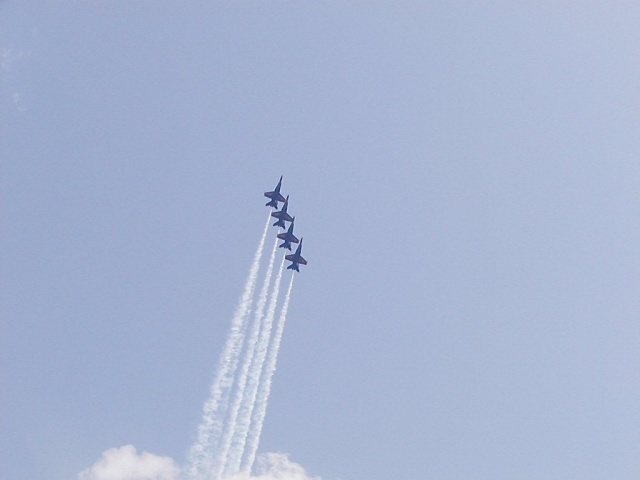 Five blue fighter jets fly in formation, leaving white smoke trails against a clear blue sky