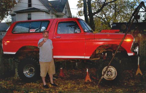 1978 Ford Bronco - A red 1978 Ford Bronco is lifted on jack stands in a yard, with a man standing beside it.