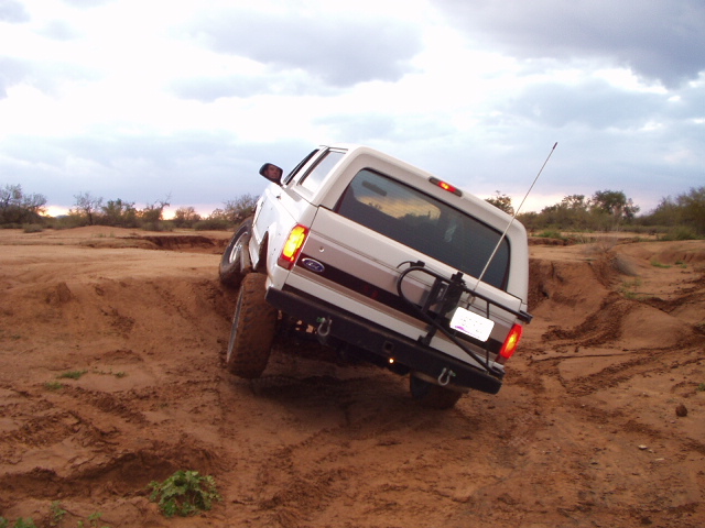 White Ford Bronco SUV tilted while climbing sandy off-road terrain, rear view