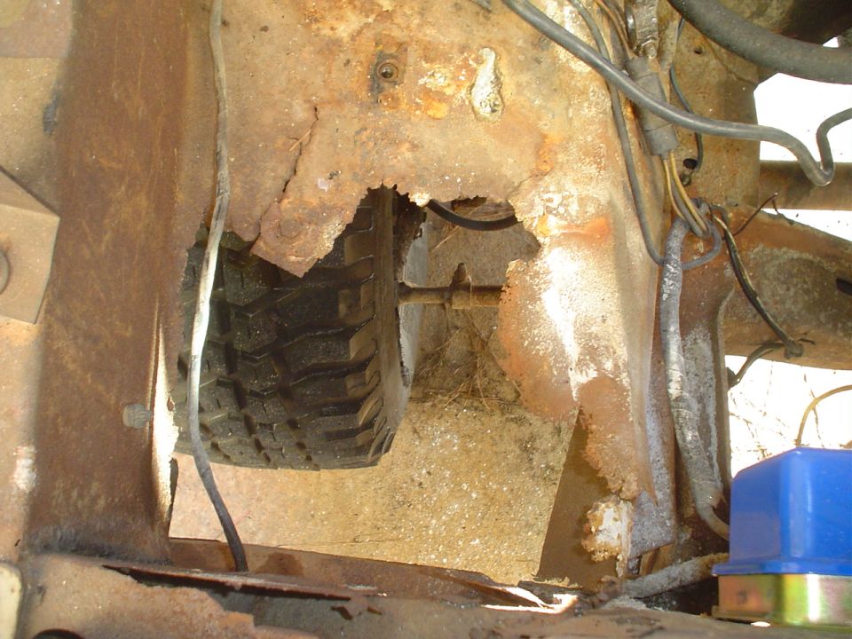 Heavily rusted vehicle frame showing a hole with a tire and suspension components visible