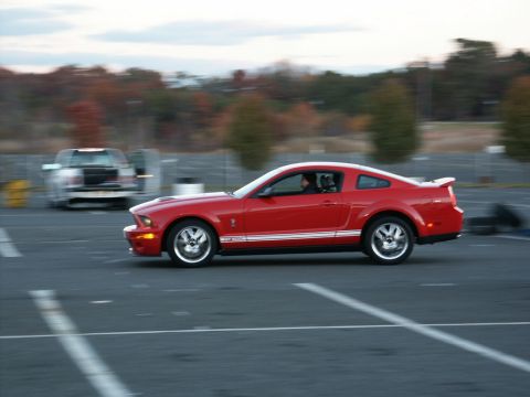 2001 Mercury Grand Marquis - A red Ford Mustang GT500 is seen in profile on a paved surface, with another vehicle blurred in the background.