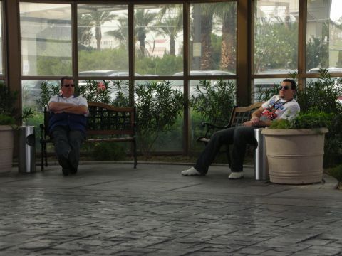 1998 Ford Crown Victoria - Two men relax on benches in a glass-walled atrium overlooking palm trees, with potted shrubs and a stone-tiled floor.