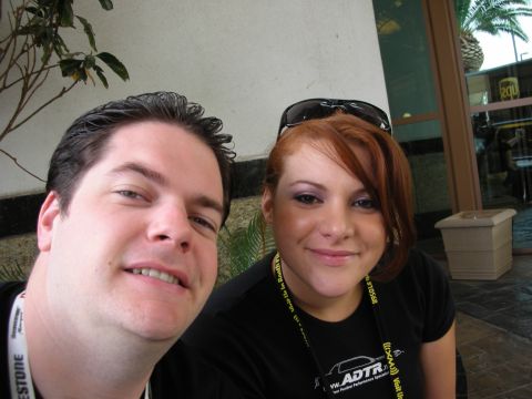 1998 Ford Crown Victoria - close selfie shows a smiling man and a red-haired woman in black shirts outdoors beside a building with greenery behind them.