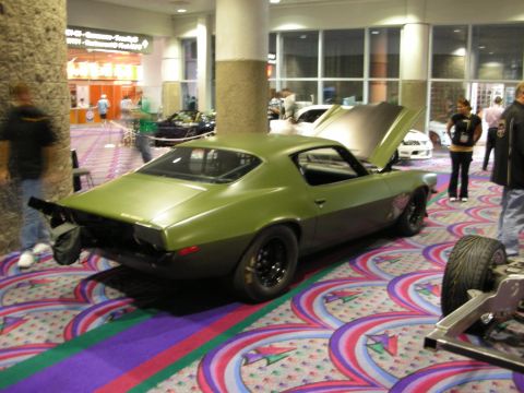 1998 Ford Crown Victoria - matte green two-door muscle car with its rear hatch open sits on colorful patterned carpet at an indoor exhibition.