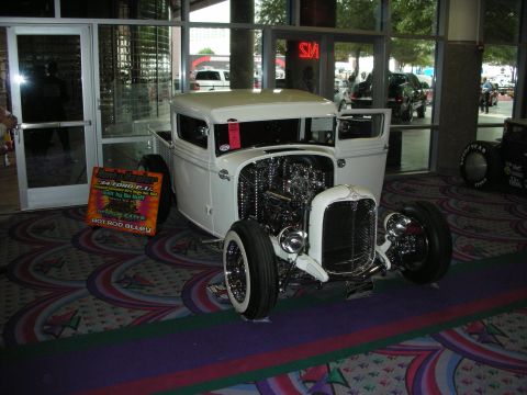 1998 Ford Crown Victoria - white chopped hot rod pickup with an exposed chromed engine and whitewall tires sits on patterned carpet inside a venue.