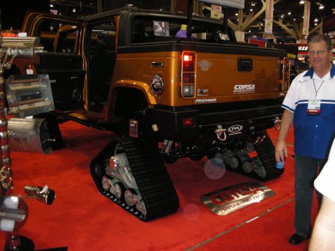 1998 Ford Crown Victoria - bronze Hummer SUV fitted with rubber tank tracks instead of wheels sits on red carpet at an indoor trade expo as a man walks past.