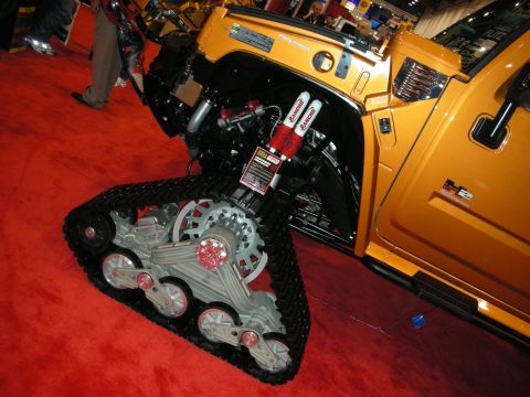 1998 Ford Crown Victoria - orange show SUV fitted with a triangular rubber track conversion and red coilover shocks in place of the front wheel, on red carpet.