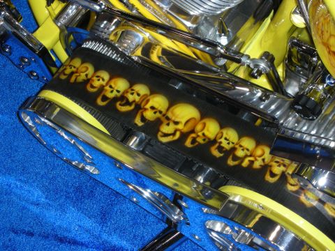 1998 Ford Crown Victoria - close-up of a chrome-and-yellow custom engine with a row of airbrushed skulls across a carbon-look panel, on blue carpet.