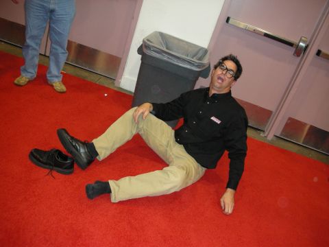 1998 Ford Crown Victoria - man in a black shirt and khaki pants reclines playfully on red carpet beside a trash can and double doors.