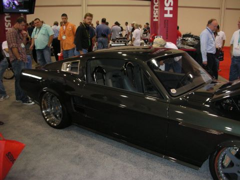 1998 Ford Crown Victoria - side profile of a lowered dark green custom Mustang fastback with chrome wheels, under a Roush banner on the show floor.