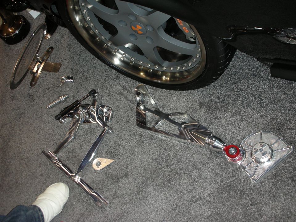 Polished chrome suspension and steering components laid out on carpet beside a car wheel