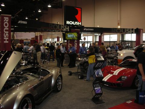 1998 Ford Crown Victoria - Busy trade-show aisle beneath ROUSH signage, with a silver roadster, red coupe and engines on display.