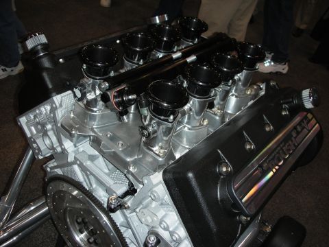 1998 Ford Crown Victoria - Roush V8 crate engine on a display stand showing black velocity-stack intakes and an exposed flywheel.