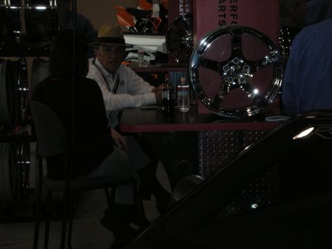 1998 Ford Crown Victoria - man in a fedora sits at a booth table next to a displayed chrome five-spoke wheel in dim lighting.