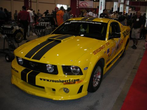 1998 Ford Crown Victoria - Yellow Roush Mustang development drag car with black stripes and roushperf.com graphics on a show floor.