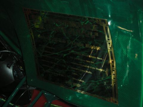 1998 Ford Crown Victoria - brass maintenance-schedule plaque with gold trim is mounted on a glossy green carbon-fiber body panel under dim light.