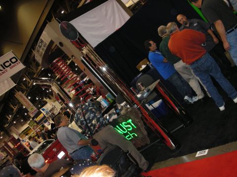 1998 Ford Crown Victoria - Visitors gather around a tall polished chrome exhaust stack with a green ENJUST sign at an indoor automotive trade show.