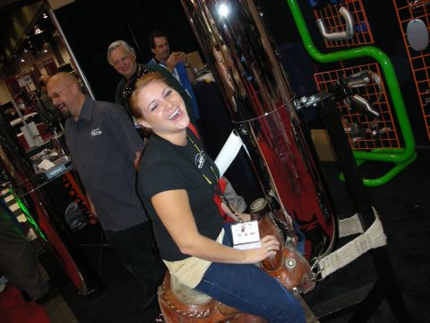 1998 Ford Crown Victoria - smiling woman in a black T-shirt sits on a leather saddle prop next to a giant chrome exhaust display at a trade show.