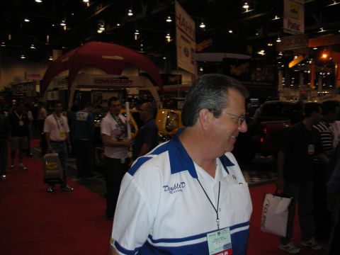 1998 Ford Crown Victoria - smiling man wearing a blue-trimmed Double D Racing polo walks a crowded car-show aisle with displays behind him.