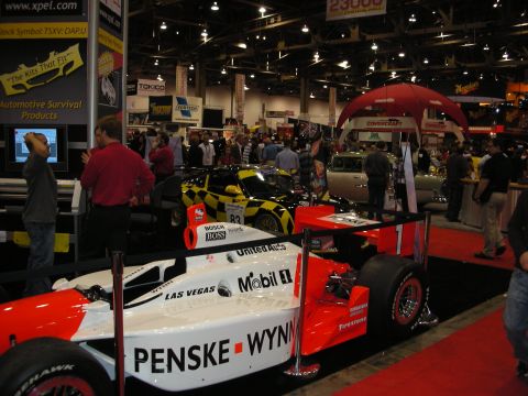 1998 Ford Crown Victoria - red and white open-wheel race car in Penske Wynn's livery sits on a display stand among booths at an indoor show.