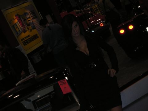 1998 Ford Crown Victoria - woman in a black dress poses at a very dim car-show booth, with taillights of display cars glowing in the background.