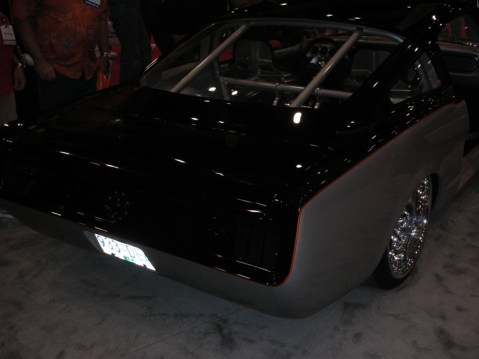 Rear quarter of a black fastback muscle car with chrome wheels and a roll cage, at an indoor show