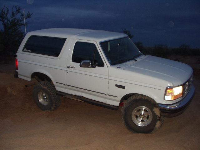 White lifted two-door Ford Bronco SUV on dirt at dusk, front 3/4 view with headlights on