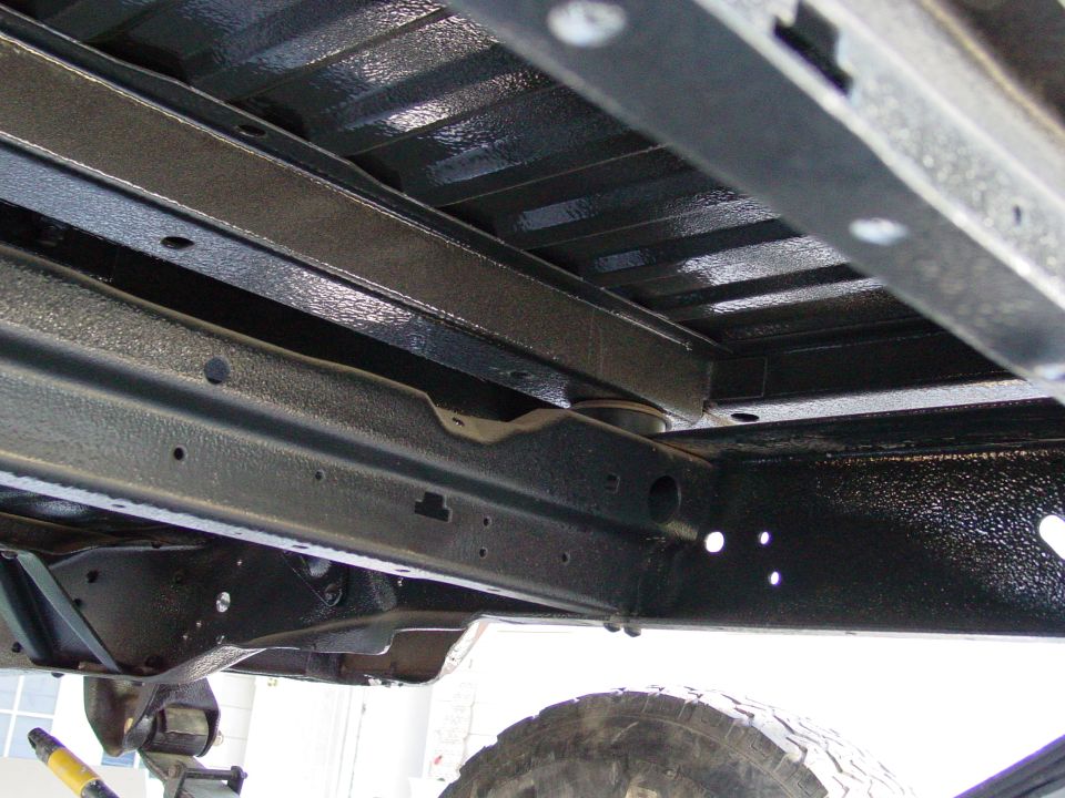 underbody of a vehicle with frame rails and crossmembers coated in black protective material, tire visible at bottom right