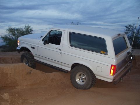 1993 Ford Bronco - White Ford Bronco with large tires navigates a dirt obstacle under a cloudy sky.