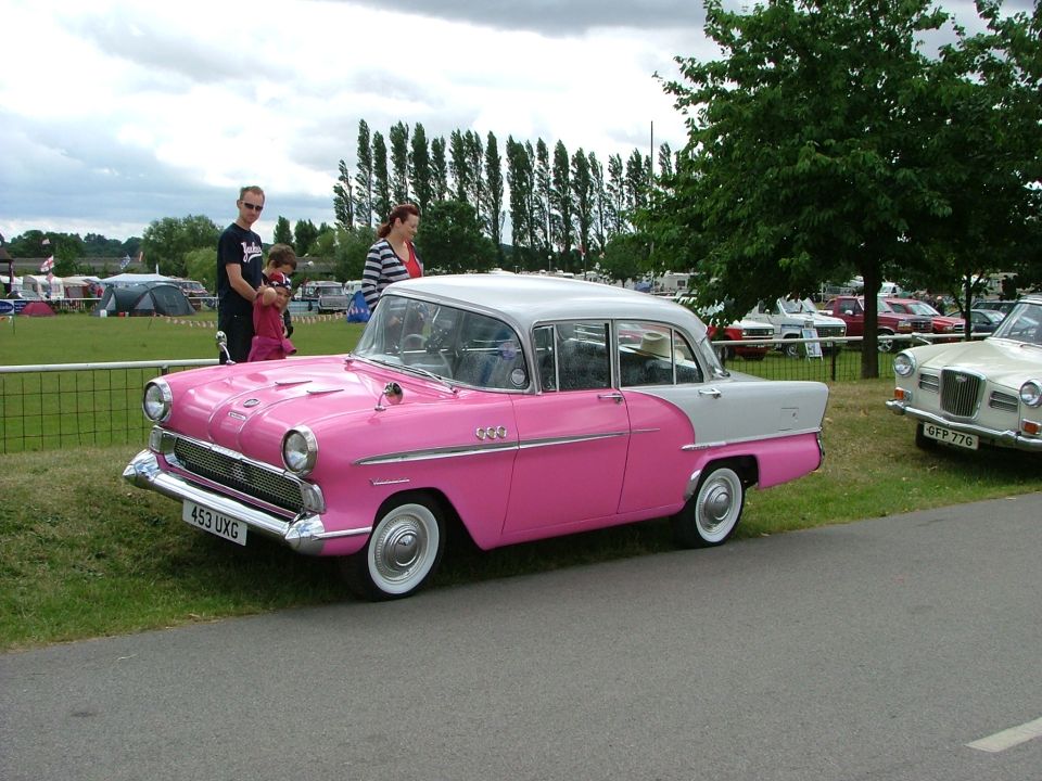 Pink and grey classic sedan parked on grass, people nearby. Another car is to the right