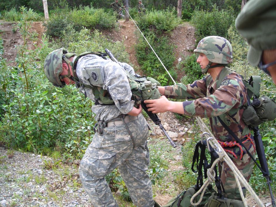 Two soldiers in camouflage uniforms and helmets practice rappelling with ropes and gear in a wooded area