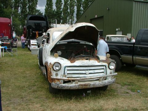 2001 Ford Fiesta - A weathered GMC truck sits in a grassy lot with its hood open, next to other vehicles and a building.
