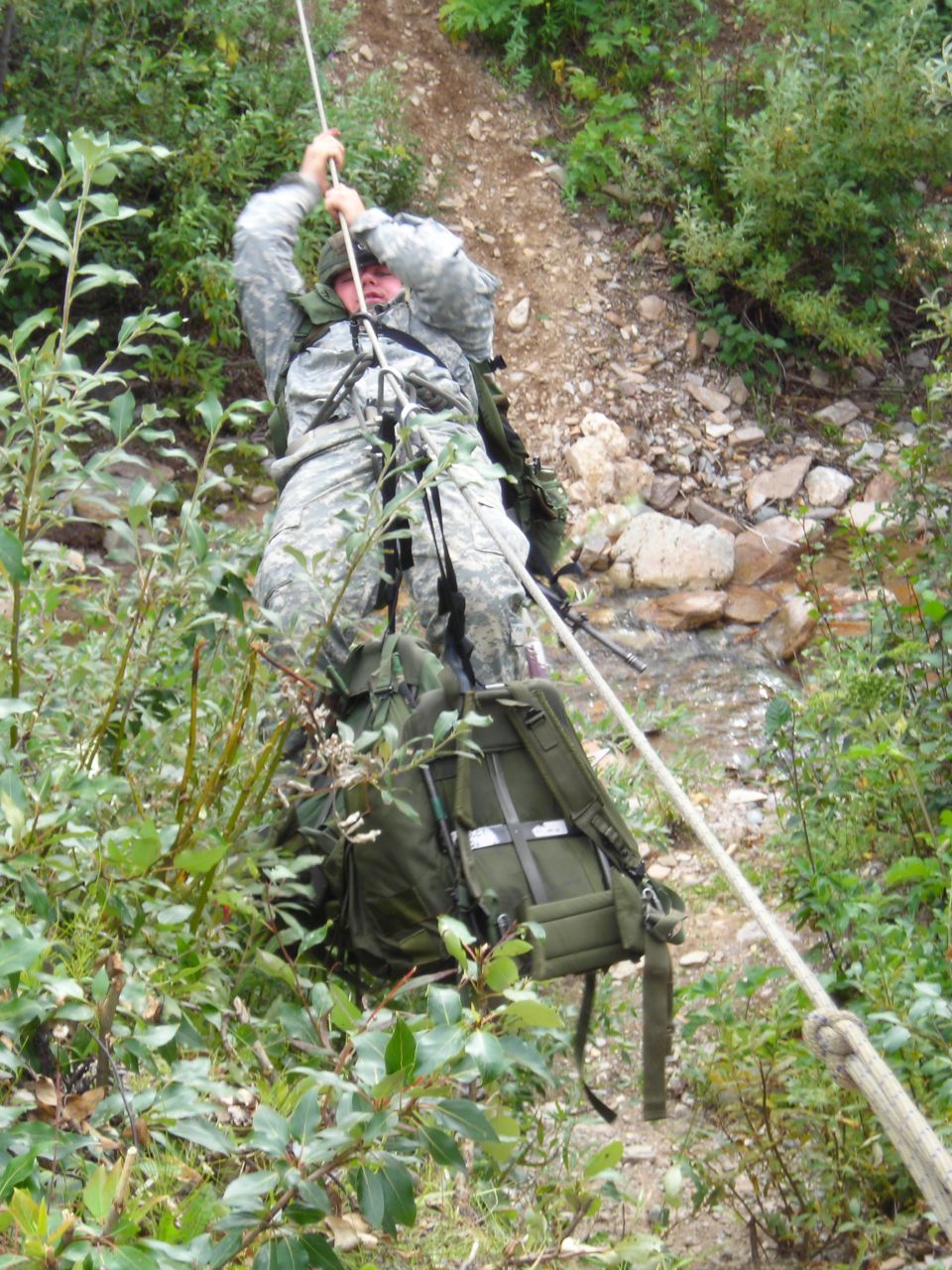 Soldier in camouflage uniform rappelling down a rocky, wooded slope using a rope