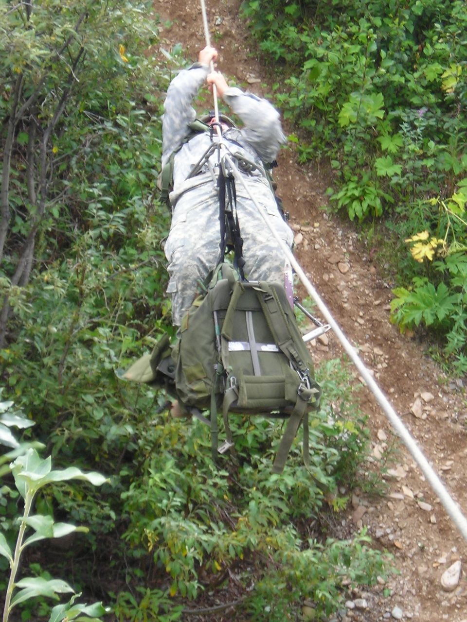 Soldier in camouflage rappelling down a steep, wooded slope using a rope, carrying a large backpack