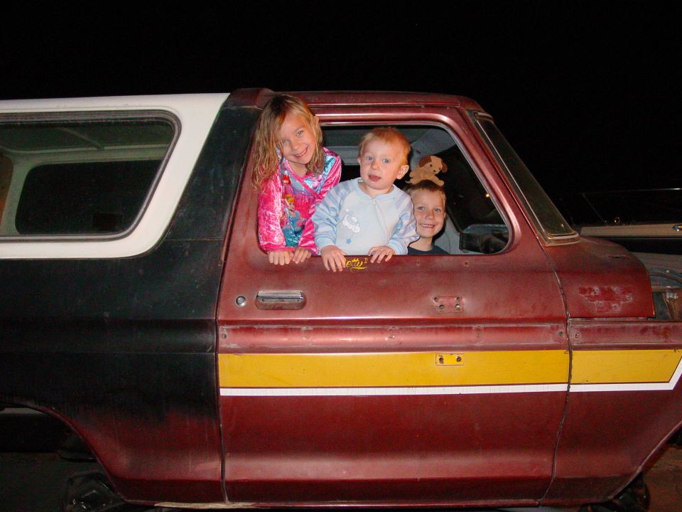 Three children look out the windows of a red and black SUV with a yellow stripe