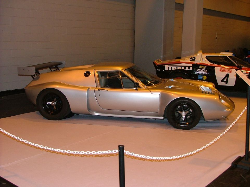 Silver sports car with a large rear spoiler and black wheels, viewed from the side