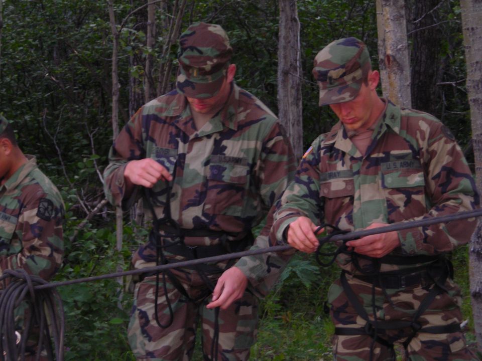 Three soldiers in camouflage uniforms practice tying knots with ropes in a wooded area