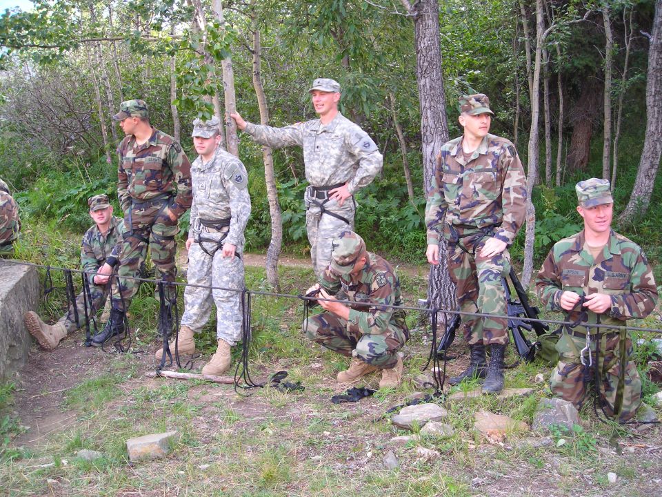 Several soldiers in camouflage uniforms stand and crouch outdoors, some wearing climbing harnesses and ropes