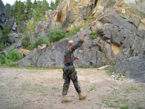 2003 Jeep TJ - A man in camouflage throws a rock at a cliff face in a wooded, rocky area.