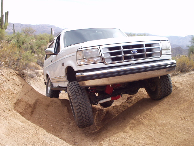 White Ford Bronco climbing dirt berm on desert trail, front 3/4 low angle