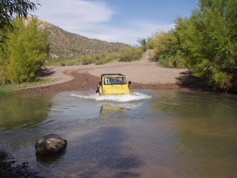 1993 Ford Bronco - A yellow Jeep navigates a shallow river crossing on a dusty trail, surrounded by arid vegetation.