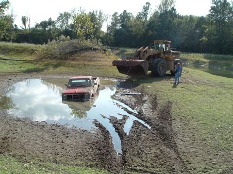 1995 Mercury Grand Marquis - A red pickup truck is submerged in a mud pit, with a front loader positioned on the bank nearby.