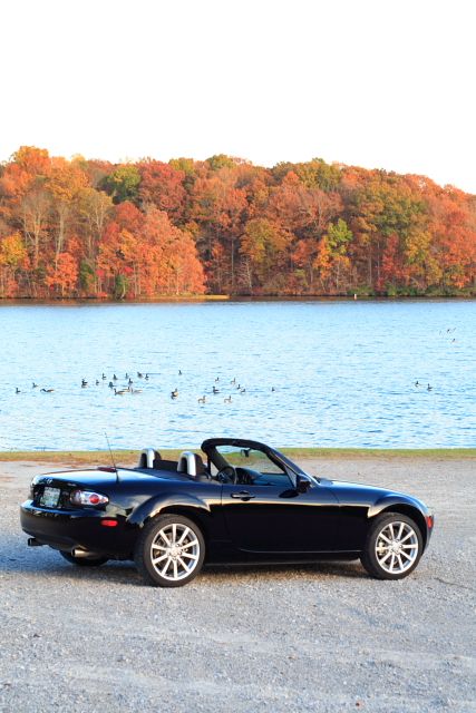 2006 Mazda Miata - A black Mazda Miata convertible sits by a lake during autumn, with trees displaying fall colors in the background.