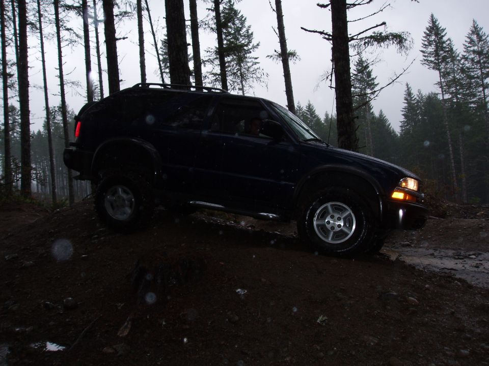 Dark blue SUV on a dirt hill in a forest, with a roof rack and aftermarket wheels