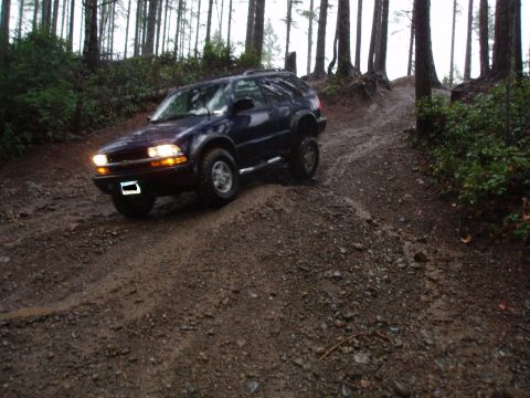 1995 Ford Bronco - A dark blue SUV navigates a challenging muddy path through a dense forest.