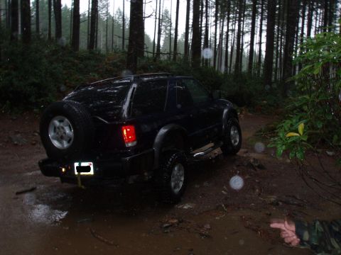 1995 Ford Bronco - A dark-colored SUV navigates a wet, muddy path through a dense forest with tall trees.
